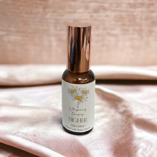 A 30ml glass bottle of 'Higher - White Quartz Crystal - Aura Spray' with a copper-colored cap, labeled with a white and yellow design, placed on a pink fabric surface.