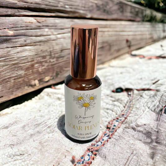 A 30ml glass bottle of 'Solar Plexus - Moonstone Crystal - Aura Spray' with a copper-colored cap, labeled with a white and green design, placed on a sandy surface with a wooden object and a multicolored rope in the background.